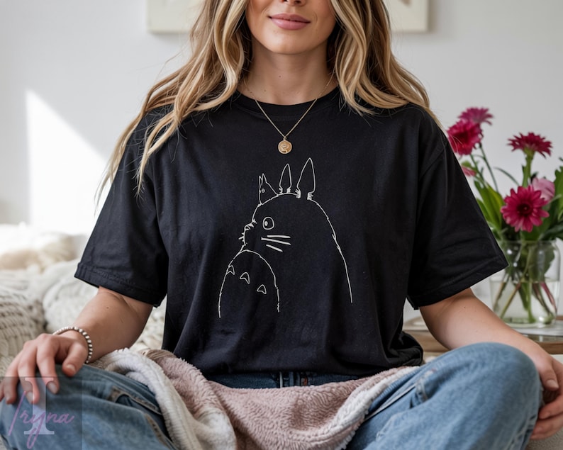 Op de afbeelding: Zwart T-shirt met een witte omtrek van twee Totoro-personages. Het ontwerp is gecentreerd op de voorkant. Het shirt is gemaakt van een zacht materiaal en heeft korte mouwen.