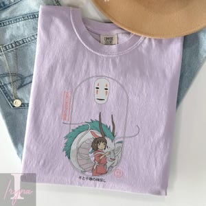 May include: A light purple t-shirt featuring a graphic from the animated film Spirited Away. The design includes the character No-Face, a white dragon, and a girl in a red dress. Japanese text is also visible.