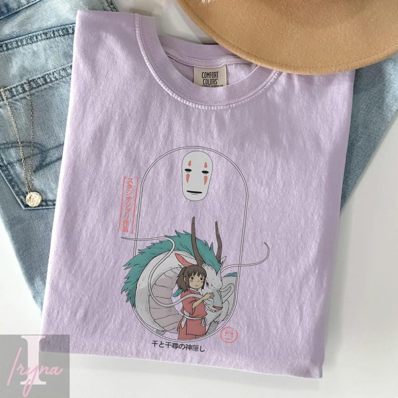 May include: A light purple t-shirt featuring a graphic from the animated film Spirited Away. The design includes the character No-Face, a white dragon, and a girl in a red dress. Japanese text is also visible.