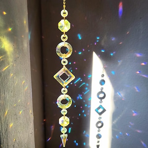 Könnte beinhalten: Ein hängender Sonnenfänger mit einer goldfarbenen Kette und verschiedenen geometrischen Formen. Der Sonnenfänger enthält eine Reihe klarer, facettierter Kristalle in Form von Kreisen, Achtecken und Rauten. Das Sonnenlicht erzeugt einen Regenbogeneffekt.