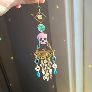 May include: A gold-colored suncatcher with a butterfly and skull design. The suncatcher features a diamond-shaped top with a butterfly, a teal-colored gem, a skull, and a gold-colored butterfly with hanging blue and pink gems.