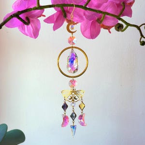 May include: A gold-colored suncatcher with a crescent moon, a large crystal teardrop, and a butterfly design. The suncatcher also features various crystal shapes and colors, including pink, purple, and yellow. The suncatcher is hanging in front of pink orchid flowers.