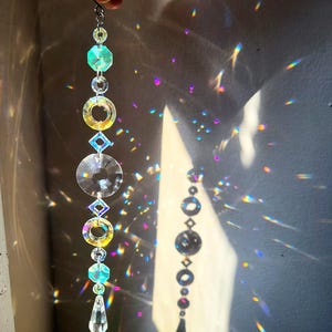 May include: A decorative crystal suncatcher with various shapes, including octagons, circles, and diamonds, strung together with a silver chain. The suncatcher casts colourful light patterns on the wall.