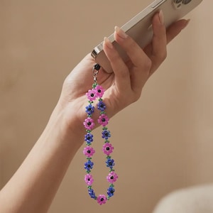 Può includere: Un cinturino per telefono con perline con un motivo floreale. Il cinturino alterna perline a forma di fiore rosa e blu con centri neri, collegate da perline verdi. Il cinturino è attaccato a un telefono argentato.