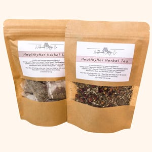 Könnte beinhalten: Zwei braune Papierbeutel mit HealthyHer Herbal Tea von Wildwood Cottage Co. Der linke Beutel enthält Teebeutel, der rechte losen Tee. Die Etiketten führen Zutaten wie Orangenschale, grüne Minze, Brennnessel, Himbeerblätter und rote Rosenblüten auf.