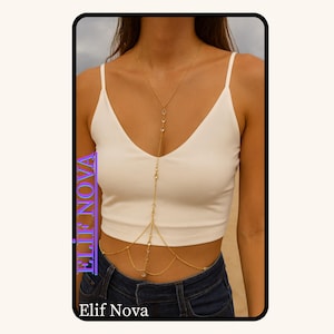 Op de afbeelding: Een goudkleurige lichaamsketting met heldere kristallen accenten. De ketting loopt van de nek, over de borst en rond de taille. De merknaam "Elif Nova" is zichtbaar in de afbeelding.