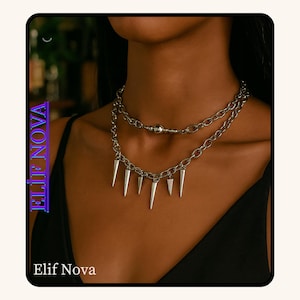 May include: Silver-toned layered necklace with a spike pendant. The necklace has two layers of chain, with a ball and chain detail on the upper layer. The lower layer features multiple cone-shaped spikes. The brand name "Elif Nova" is displayed in the image.