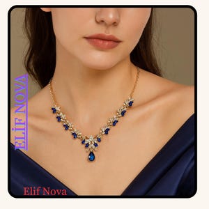 May include: A gold-toned necklace with blue and clear gemstones. The necklace features a central teardrop-shaped blue gem and a floral design. The brand name "Elif Nova" is visible in the image.