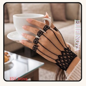May include: Black hand chain jewelry with intricate lace-like designs on the fingers and wrist. Delicate chains connect the finger accents to the wrist cuff. The image also includes a white teacup and saucer. The text "Elif Nova" is visible.