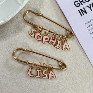 Peut inclure: Deux épingles de sûreté dorées avec des breloques en forme de lettres roses émaillées épelant "SOPHIA" et "LISA". Les épingles sont posées sur une surface neutre, avec une soucoupe blanche et un papier avec du texte en arrière-plan.