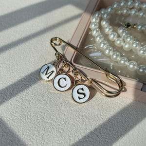 Może przedstawiać: Złota agrafka z trzema białymi, okrągłymi zawieszkami z czarnymi literami M, C i S. Agrafka leży obok naszyjnika z pereł w różowym pudełku na biżuterię. Ozdobny dodatek.