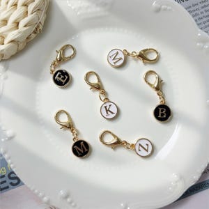 Könnte beinhalten: Goldfarbene Metallanhänger mit Karabinerverschlüssen, jeweils mit einem runden Anhänger mit einem Buchstaben. Die Anhänger sind entweder schwarz mit weißem Buchstaben oder weiß mit schwarzem Buchstaben. Die sichtbaren Buchstaben sind E, M, K, B und Z.