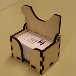 May include: A wooden business card holder with a hinged lid. The holder is made of light-colored wood and contains a stack of white business cards. The cards have text and a logo printed on them.