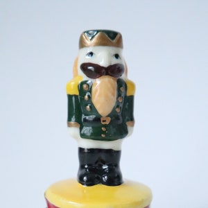 May include: A porcelain nutcracker figurine with a green and gold uniform, a white face, and a black mustache. The figurine is standing on a red and yellow drum-shaped box.