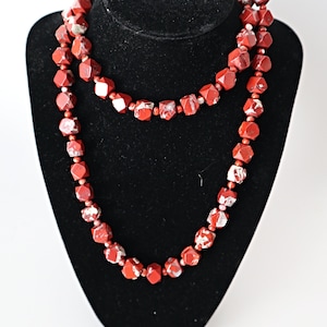 May include: A red and white gemstone necklace with faceted beads. The necklace is displayed on a black velvet stand.