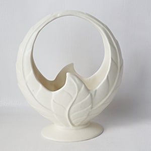 May include: Off-white ceramic basket with a large handle. The basket is shaped like a leaf, with detailed veining. The base is round and the handle curves gracefully. A decorative piece for home decor.