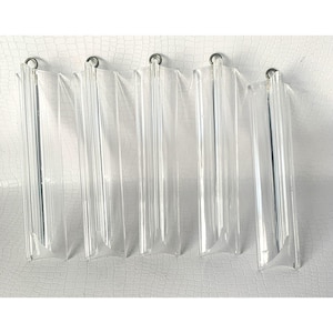 Puede incluir: Cinco prismas de araña de acrílico transparente y rectangular con anillos de metal plateado en la parte superior. Los prismas están orientados verticalmente y tienen un diseño acanalado. El fondo es una superficie blanca texturizada.