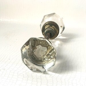 May include: Two clear, faceted glass doorknobs with brass fittings and a metal rod. The knobs are octagonal and show signs of age. The background is a white, textured surface.