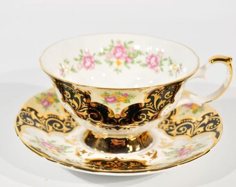 May include: A white teacup and saucer with a black and gold floral pattern. The cup has a delicate handle and the saucer has a scalloped edge.