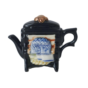 Puede incluir: Una pequeña tetera de cerámica negra con un diseño detallado. La tetera presenta un pequeño acento decorativo de color cobre en la tapa y un relieve detallado de una estructura azul y blanca en la parte delantera. El asa y el pico son curvos.