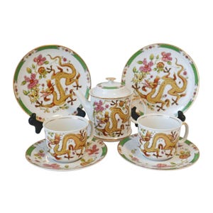 Peut inclure: Un service à thé en porcelaine blanche avec un motif de dragon. L'ensemble comprend une théière, deux tasses et deux soucoupes. Le dragon est représenté en or et rouge, avec des accents floraux verts et roses.