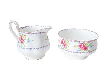 Può includere: Un set di lattiera e zuccheriera in porcellana bianca. Il set presenta un motivo floreale con fiori rosa, blu e verdi. I bordi sono rifiniti con oro. La lattiera ha un manico e un beccuccio. Lo zuccheriera ha un'ampia apertura.