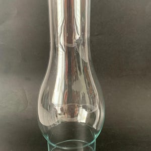 May include: A clear glass lamp chimney with a bulbous base that tapers into a tall, slender cylindrical neck. The glass is transparent and smooth, featuring a wide circular opening at the bottom and a smaller circular opening at the top.