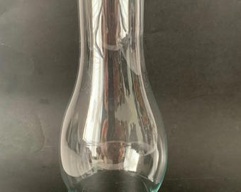 May include: A clear glass lamp chimney with a bulbous base that tapers into a tall, slender cylindrical neck. The glass is transparent and smooth, featuring a wide circular opening at the bottom and a smaller circular opening at the top.