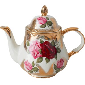 Peut inclure: Une théière en porcelaine blanche avec une bordure dorée et un motif floral composé de roses roses et rouges. La théière a un couvercle avec un bouton doré.