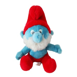 May include: A blue plush toy of a Smurf character with a white beard, red hat, and a white and blue outfit. The Smurf is sitting with its legs crossed.