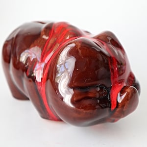 Könnte beinhalten: Ein braunes Keramik-Sparschwein mit roter Glasur. Das Sparschwein liegt auf der Seite mit der Schnauze zur Kamera.