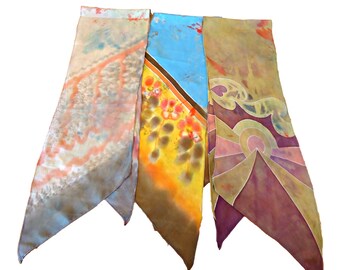 May include: Three silk scarves with abstract designs. One scarf features a light blue and peach pattern, another has a blue and yellow design with orange accents, and the third has a purple and gold design.