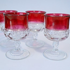 May include: Four clear glass goblets with ruby red rims. The lower half of each glass features a decorative, etched pattern. The goblets have a classic design, with a round base and a stem.