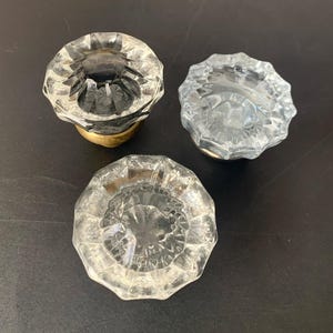 May include: Three clear glass knobs with a faceted, multi-sided design. Each knob features a circular top with a starburst pattern radiating from the center and a metallic base. The glass is transparent and reflects light across its textured surface.