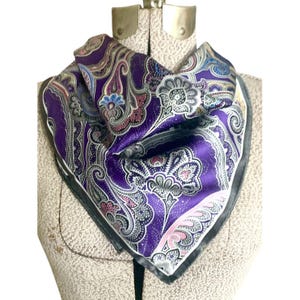 May include: A square silk scarf with a paisley pattern in shades of purple, pink, blue, and silver. The scarf has a black border and is displayed on a mannequin. The scarf is a fashion accessory.