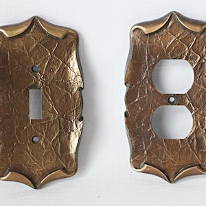 May include: Two decorative wall plates in a gold finish with a textured surface. One plate has a single light switch opening and the other has a double outlet opening.