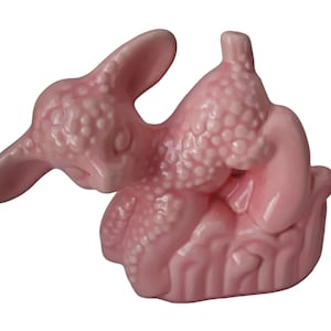 May include: A pink ceramic figurine of a sleeping lamb. The lamb is lying on its side with its head resting on its front legs. The figurine is detailed with a textured surface and has a small, pink base.