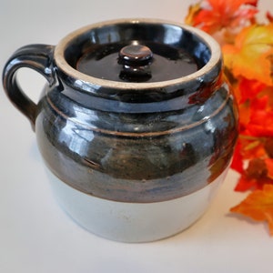 May include: A brown and white glazed ceramic crock with a lid. The crock has a dark brown glaze with a white band around the bottom. The lid has a dark brown glaze with a small knob on top.