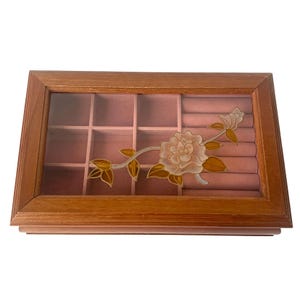 Peut inclure: Boîte à bijoux en bois avec un dessus en verre transparent et un intérieur en velours rose. La boîte présente un motif floral avec une rose et des feuilles dans les tons de rose, blanc et marron. La boîte est divisée en compartiments.