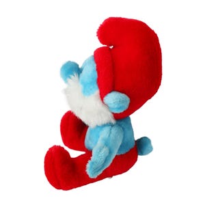 May include: A blue and red plush toy of a Smurf character. The Smurf is sitting with its legs crossed and has a white beard. The Smurf is wearing a red hat.