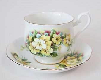 May include: A white teacup and saucer set with a floral design featuring yellow primroses and green leaves. The cup has a gold rim and the saucer has a scalloped edge.