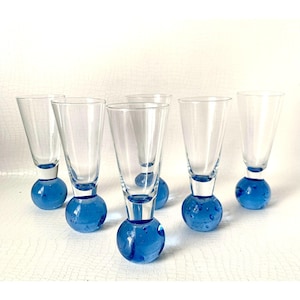 May include: A set of six clear glass flutes, each featuring a distinct spherical base in a vibrant cobalt blue colour. The glass bowls are tall and slender, tapering slightly towards the bottom where they connect to the solid, bubble-filled blue glass spheres.