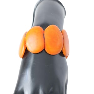 Könnte beinhalten: Ein orangefarbenes Armband mit großen, ovalen Perlen. Das Armband besteht aus glänzendem Material und wird auf einem schwarzen Mannequin-Hand gezeigt. Die Perlen sind in einem leuchtenden Orange gehalten, was es zu einem auffälligen Accessoire macht.