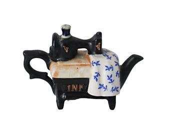 May include: A ceramic teapot in the shape of a vintage sewing machine. The teapot is black with gold detailing and a white and blue floral pattern. The base of the sewing machine reads "INF".