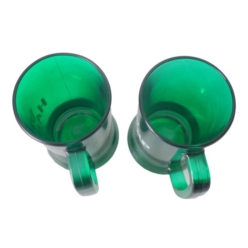 May include: Two green plastic mugs with black rims and handles are shown against a white backdrop. The mugs are transparent, revealing their interiors, and have a slightly tapered shape. The word "AH" is visible on the side of one mug.