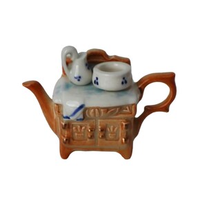 Peut inclure: Petite théière en céramique en forme de cuisinière, marron et crème. Elle comprend un bec verseur, une anse, une petite cruche et une tasse. La cuisinière présente un design détaillé avec des accents dorés. Objet décoratif charmant.