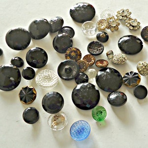 May include: A collection of vintage buttons in various colors and styles, including black, gold, clear, and green. The buttons are made of glass, metal, and plastic. Some buttons have intricate designs, while others are plain.