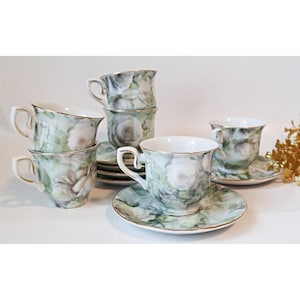 May include: Set of six white porcelain teacups and saucers with a floral design featuring white roses and green leaves. The cups and saucers have a gold trim.