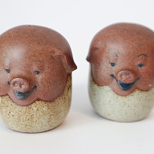 Może przedstawiać: Zestaw dwóch brązowych ceramicznych solniczek i pieprzniczek w kształcie świń z uśmiechającymi się twarzami. Solniczki mają brązową glazurę w kropki i białą glazurę na spodzie.