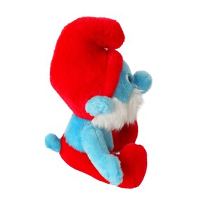 May include: A blue plush Smurf toy with a red hat and white beard. The Smurf is sitting with its legs crossed and its arms folded.